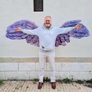 photographie d'un homme très souriant posant devant des ailes bleues de la victoire dessinées par Karine Corbier collées sur le mur de la rue
