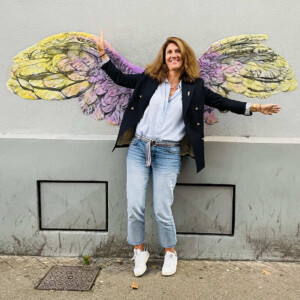 photographie d'une Jeune femme très dynamique posant devant des ailes de la victoire dessinées par Karine Corbier collées sur le mur de la rue