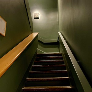 Grand escalier sombre vert au sommet duquel est accroché un tout petit tableau de 30 par 30 cm représentant un camouflage vert