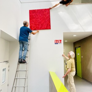 Accrochage d'un monochrome rouge de Karine Corbier en hauteur par deux assistants techniques Une personne âgée regarde la scène
