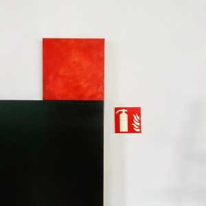 Peinture monochrome rouge de Karine Corbier posé sur une porte noire à côté d'un pictogramme incendie rouge
